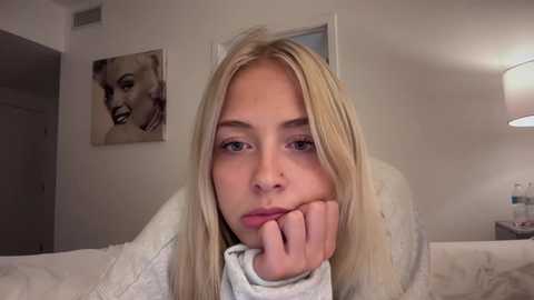 Media: Video of a blonde woman with fair skin, resting her chin on her hand, looking contemplative. Background features a white wall with a Marilyn Monroe portrait and a bedside lamp.