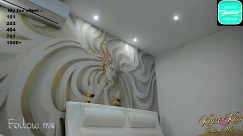 Media: Video of a modern, minimalist room with a large, intricate white mural featuring a stylized, ethereal female figure on the wall. The room has a plush white sofa, recessed lighting, and a tropical-themed air conditioner.