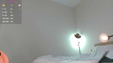 Media: A video of a minimalist bedroom featuring a bed with white sheets, a bedside lamp with a round bulb, and a large, bright studio light on the wall. The room is sparsely decorated with beige walls.