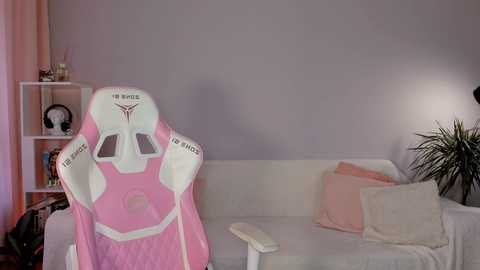 Media: Video of a pink gaming chair with \"I AM THE MIGHTY\" and \"TOO MUCH\" emblems, positioned in a minimalist room with a white couch, a green plant, and a soft pink curtain.