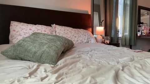 Media: Video of a cozy bedroom with a white duvet, green pillow, floral-patterned pillows, dark wooden headboard, lamp, and window with green curtains.