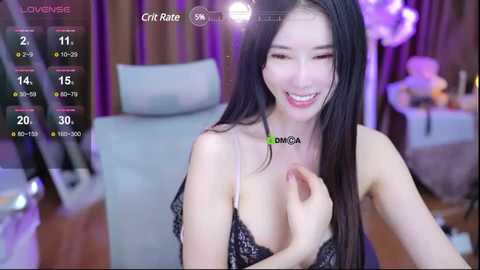 Media: Video of an East Asian woman with long black hair, wearing a black lace bra, smiling while holding a phone in her left hand. Background shows a dimly lit room with a blue sofa.