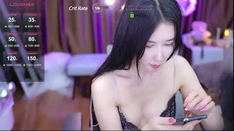 Media: Video of a slender East Asian woman with long black hair, wearing a black lace bra, focused on a smartphone in a dimly lit room.