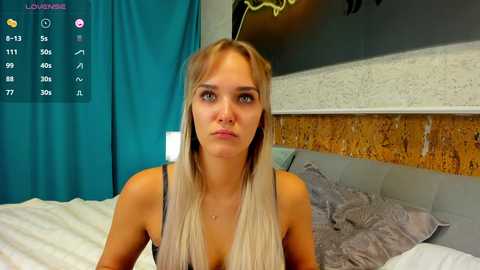 Media: Video of a blonde woman with straight hair, wearing a gray tank top, sitting on a bed in a modern bedroom with a teal curtain, beige wall, and wooden headboard.