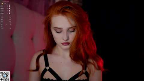 Media: A photograph of a pale-skinned, red-haired woman with long, wavy hair, wearing a black strappy lingerie top, set against a dimly lit, dark background with red lighting. She appears contemplative and introspective.