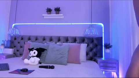 Media: A video of a modern, purple-lit bedroom with a tufted gray headboard, two potted plants, white shelves, and two glass pendant lights. A black cat toy rests on a neatly made bed.