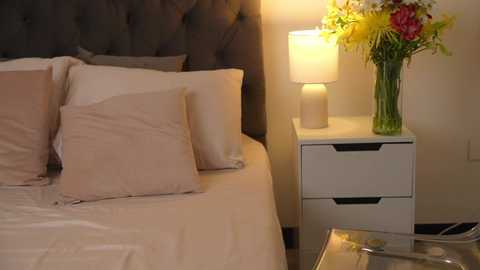 Media: Video of a cozy bedroom with a tufted dark gray headboard, beige bedding, two pillows, a nightstand with a white lamp and vibrant flower arrangement, and a sleek, modern bedside table.