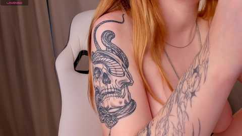 Media: Video of a fair-skinned woman with long red hair, showcasing a detailed black snake tattoo on her left shoulder, and intricate floral tattoo on her right arm. She wears a thin necklace and is seated on a white chair against a beige curtain backdrop.