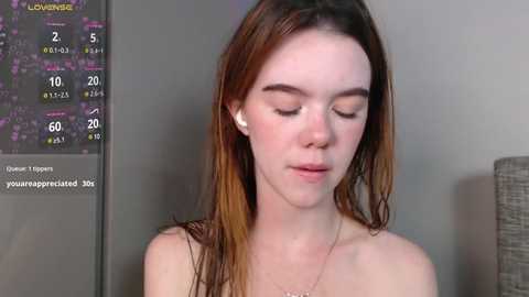 Media: Video of a fair-skinned young woman with straight, brown hair, wearing white earbuds, in a neutral expression, against a gray wall with a clock and calendar.