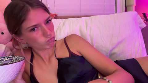 Media: Video of a young woman with light skin, brown hair, and large hoop earrings, lying on a bed with white pillows, wearing a black satin camisole, looking sultry.