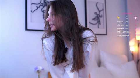 Media: A photograph of a young Caucasian woman with long brown hair, wearing a white shirt, in a modern bedroom with minimalist decor.