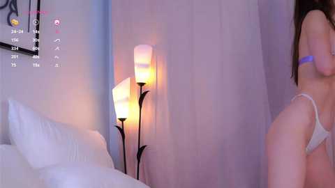 Media: A photograph of a woman in a white lace thong standing in a dimly lit bedroom with white sheets, a tall floor lamp, and a soft pink glow.