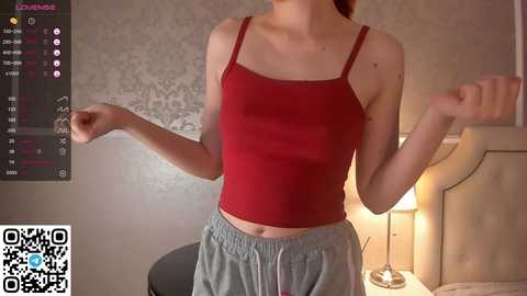 Media: Video of a slim, fair-skinned woman with red hair, wearing a red spaghetti strap crop top and gray sweatpants, in a modern bedroom with a patterned wallpaper, a lamp, and a QR code.