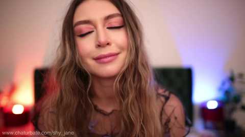 Media: Video of a young woman with long, wavy brown hair, wearing a sheer black lace top, smiling with closed eyes, set against a softly lit, blurred background.