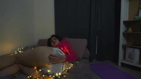 Media: Video of a woman with curly hair, wearing a red sweater, lying on a bed with string lights, in a dimly lit room.