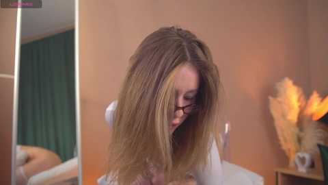 Media: Video of a woman with long, blonde hair styled in a messy, voluminous cut. She is sitting in a modern bathroom with beige walls, a green shower curtain, and a lit candle on the right.