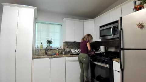 Media: A video of a modern kitchen featuring a woman with curly hair, wearing a maroon top and green pants, cooking on a stove. White cabinets, granite countertops, and a microwave are visible.