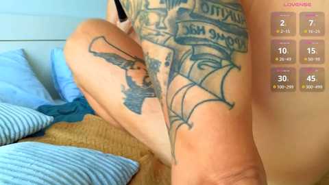 Media: Video of a person's upper thigh with a large, detailed tattoo featuring a bird and wings, lying on a bed with striped pillows and a woven blanket.