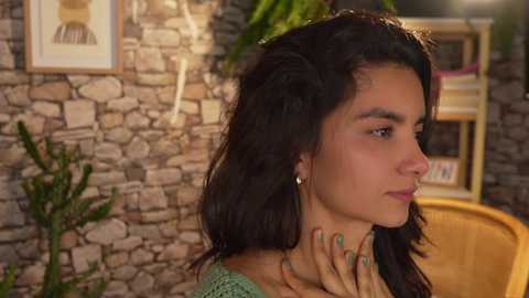 Media: Video of a young woman with dark hair, olive skin, and light makeup, holding her neck in a cozy room with a stone wall, plants, and wooden furniture.