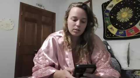 Media: A video shows a young woman with long, wavy brown hair in a pink satin robe, focused on her smartphone. She is in a dimly lit room with a wooden door, a tapestry of the zodiac, and a white wall.