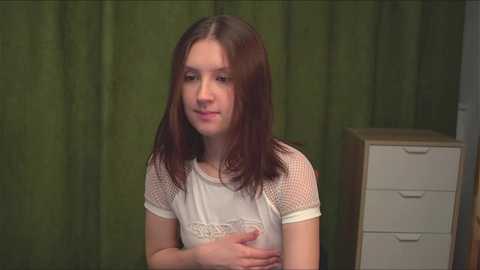 Media: Video of a young girl with long brown hair, wearing a white lace top, standing against a green curtain backdrop, with a white dresser to the right.