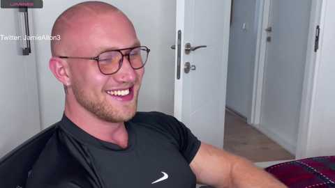 Media: Video of a smiling bald man with glasses, wearing a black Nike t-shirt, seated indoors. Background features a white door and tiled floor.