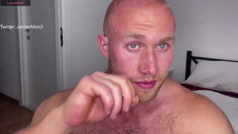 Media: Video of a muscular, bald, light-skinned man with a beard, wearing a nose ring, touching his chin, in a bedroom with a bed and white walls.