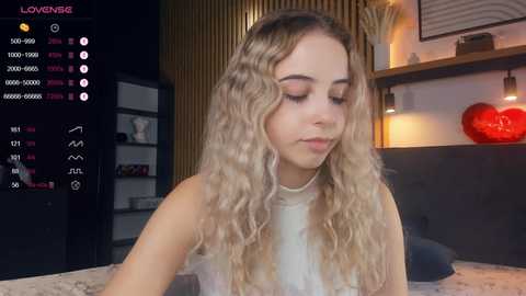 Media: Video of a young woman with long, wavy blonde hair in a white top, sitting on a bed. Background features a wooden wall, a red heart decoration, and a dimly lit room.