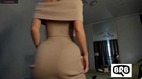 Media: Video of a woman in beige ribbed dress, viewed from behind, showing off her curvaceous buttocks. Background shows a dimly lit room with a bed and a mirror.