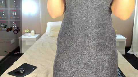 Media: A video of a woman in a gray, textured dress standing in a modern bedroom. The background features a white bed, white nightstands, and a window with white curtains.