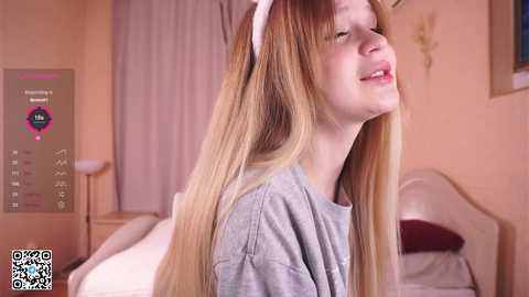 Media: Video of a smiling, fair-skinned girl with long blonde hair, wearing a grey shirt, in a pink bedroom.