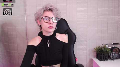 Media: Video of a light-skinned person with short, platinum blonde hair, wearing black glasses, a black crop top, and sitting in a black gaming chair, against a light, textured background.