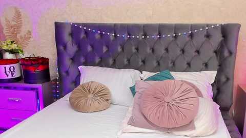 Media: Video of a chic, modern bedroom with a gray tufted headboard, white linens, two decorative pillows, and fairy lights. The background features a beige textured wall and a nightstand with a vase of flowers and a red box.