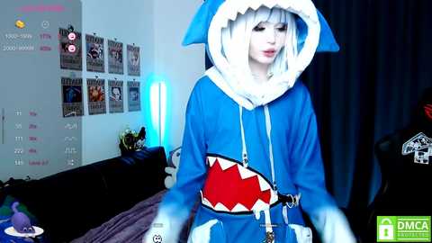 Media: Video of a pale-skinned woman in a blue shark onesie with a white hood and a red shark mouth, standing in a dimly lit room with posters and a bed.