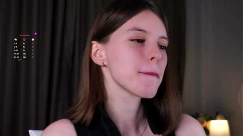 Media: Video of a young Caucasian woman with straight brown hair, wearing a black sleeveless top, looking contemplative. Background features dark curtains and a lamp.