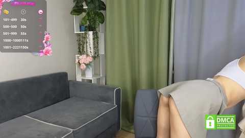 Media: Video of a woman in a grey skirt bending over a grey couch, with a green curtain and plant in the background, overlaying a screen with a pink floral design.