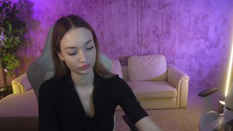 Media: Video of a young woman with straight brown hair, wearing a black top, seated in a modern, beige leather armchair. Background includes a purple-lit textured wall and a green potted plant.