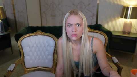 Media: A video of a blonde woman with fair skin and large eyes, wearing a teal bra, sitting on a vintage green chair with white upholstery. The background features a lamp, a floral-patterned wall, and a dark wooden table.