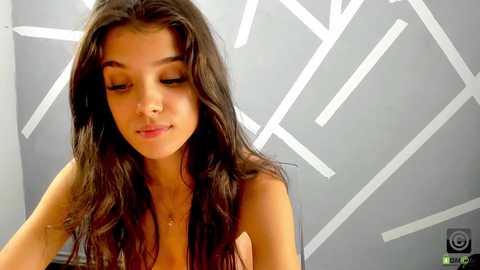 Media: A video of a young woman with long, dark hair, closed eyes, and a subtle smile, standing indoors against a modern, grey wall with white geometric patterns.