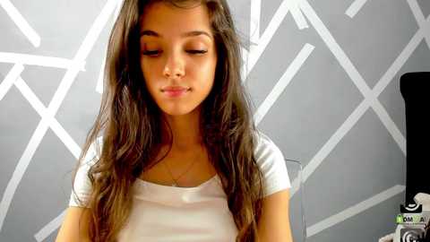 Media: Video of a young Latina girl with long, wavy dark brown hair, wearing a white T-shirt, against a gray geometric-patterned wall. She has a neutral expression.