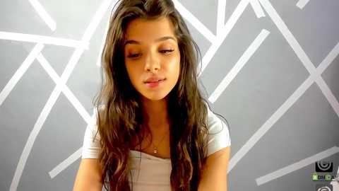 Media: Video of a young Latina woman with long, wavy dark brown hair, wearing a white short-sleeved top. She has a light skin tone and is looking down thoughtfully. Background features a geometric, white-patterned wall.