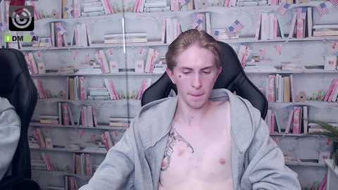 Media: Video of a shirtless young man with a slender build, pale skin, and shoulder-length blonde hair, sitting in a black office chair. He wears an open grey jacket, revealing a tattoo on his chest. The background features a bookshelf filled with books and a small American flag.