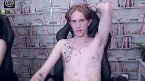 Media: Video of a slender, pale-skinned, androgynous man with long, straight, light brown hair, seated in a black gaming chair, wearing a black shirt, with tattoos on his chest and arms, in a room with bookshelves filled with colorful books.