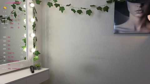 Media: Video of a small, cozy makeup room with a white wall adorned with a calendar, green ivy garland, and a framed portrait. A white vanity table holds a plant and a makeup brush holder.