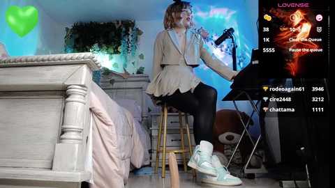 Media: Video of a woman in a beige jacket, black leggings, and sneakers playing a guitar on a stool in a cozy, dimly-lit room with a bed, green heart, and musical icons in the corner.