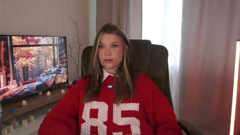 Media: Video of a young woman with light skin, long brown hair, wearing a red sweater with the number \"85\" on it, seated in a dark brown office chair, with a flat-screen TV showing autumnal trees in the background.