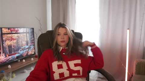 Media: Video of a young woman with light skin, long brown hair, and a red \"BRE\" sweater, sitting in a brown armchair in a cozy, brightly lit room with a TV showing a fall forest scene.