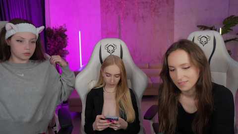 Media: Video of three young women, one in a grey sweatshirt, another in a black top, and the third in a pink sweater, seated in white gaming chairs with cat ears, in a dimly lit room with purple lighting and a potted plant.