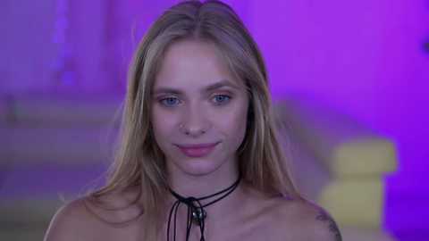 Media: Video of a young, fair-skinned, blonde woman with blue eyes and a nose piercing. She has a tattoo on her left shoulder. The background is purple and blurry, possibly indicating an indoor setting.