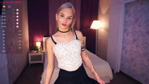 Media: Video of a young, light-skinned woman with blonde hair, wearing a white lace top and black skirt, sitting on a bed in a dimly-lit, purple-themed room.